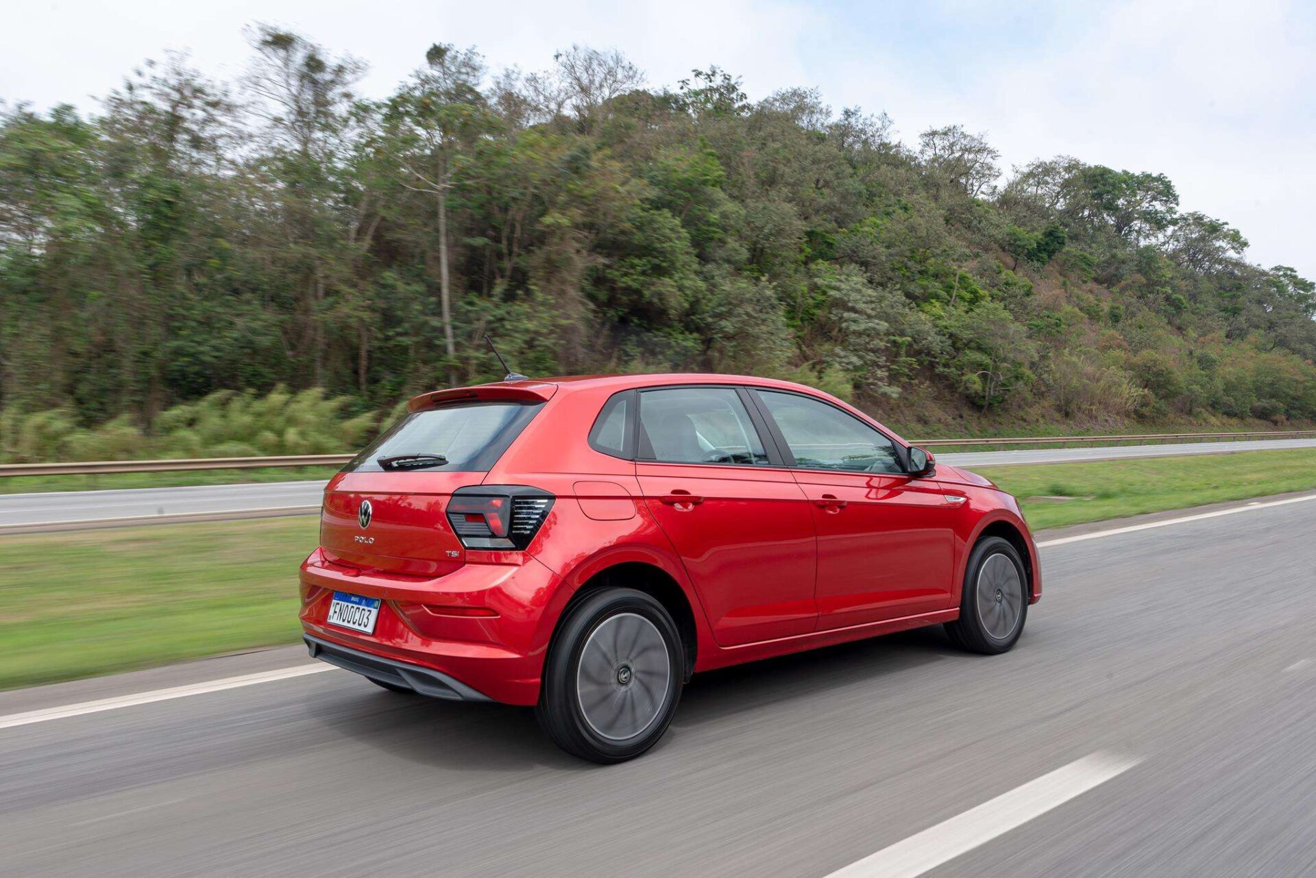 Um carro hatchback vermelho em movimento em uma estrada, cercado por uma vegetação ao fundo.