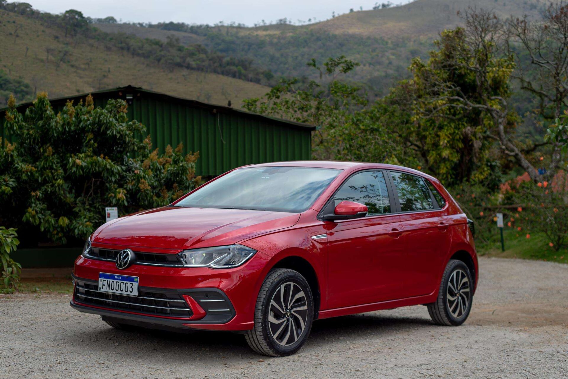 A red Volkswagen car parked on a gravel surface with green trees and a hillside in the background.