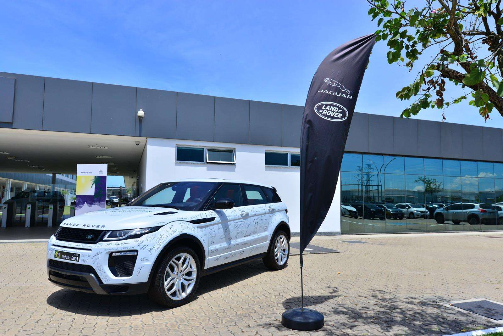 A white Range Rover Evoque with signatures on its surface parked outside a dealership, with a black flag displaying the Jaguar and Land Rover logos in the foreground.