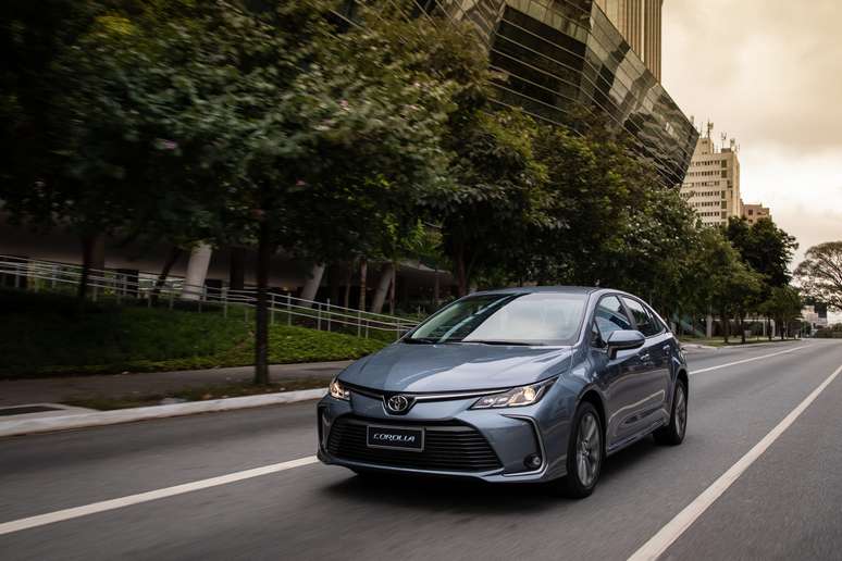 A blue Toyota Corolla driving on a city street with trees and modern buildings in the background.