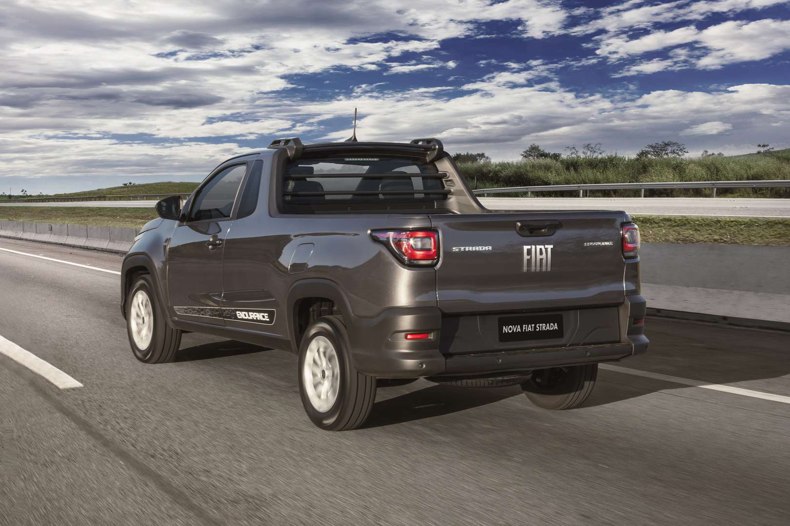 A gray Fiat Strada pickup truck driving along a highway under a partly cloudy sky.