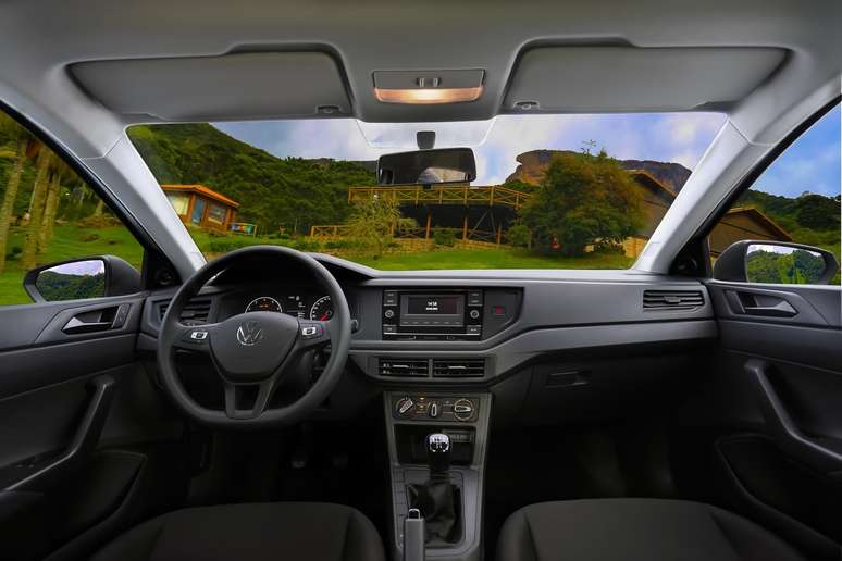 Interior view of a modern car showcasing the steering wheel, dashboard with infotainment system, gear shift, and large front windshield reflecting a scenic outdoor landscape.