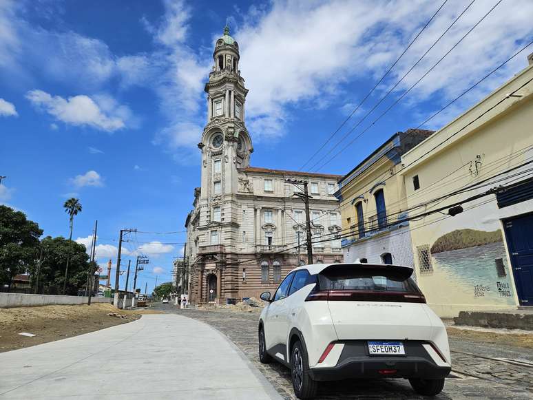 Um carro branco estacionado em uma rua de paralelepípedos, com um edifício histórico ao fundo sob um céu azul com nuvens brancas.