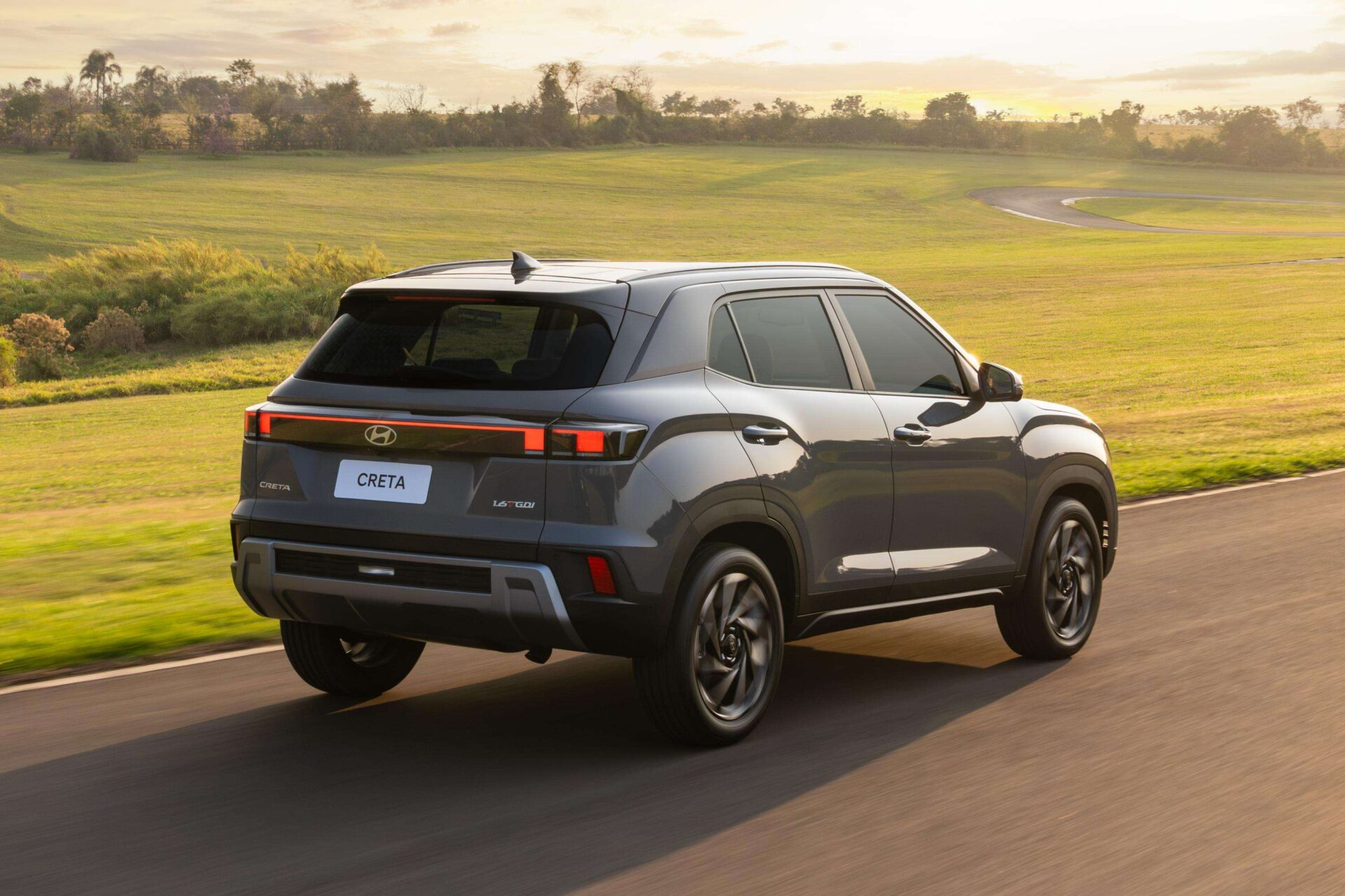 A Hyundai Creta SUV driving on a winding road with lush green fields and trees in the background during sunset.
