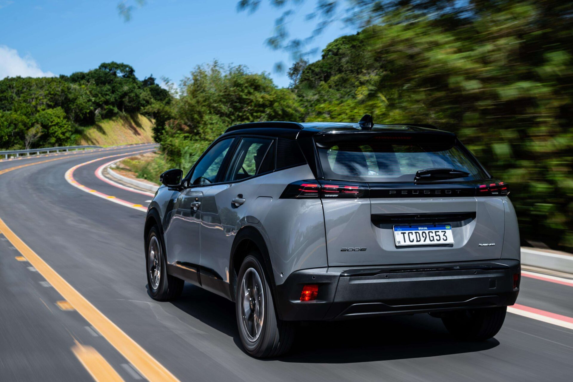 A silver Peugeot 3008 SUV driving on a curved road surrounded by greenery under a clear blue sky.