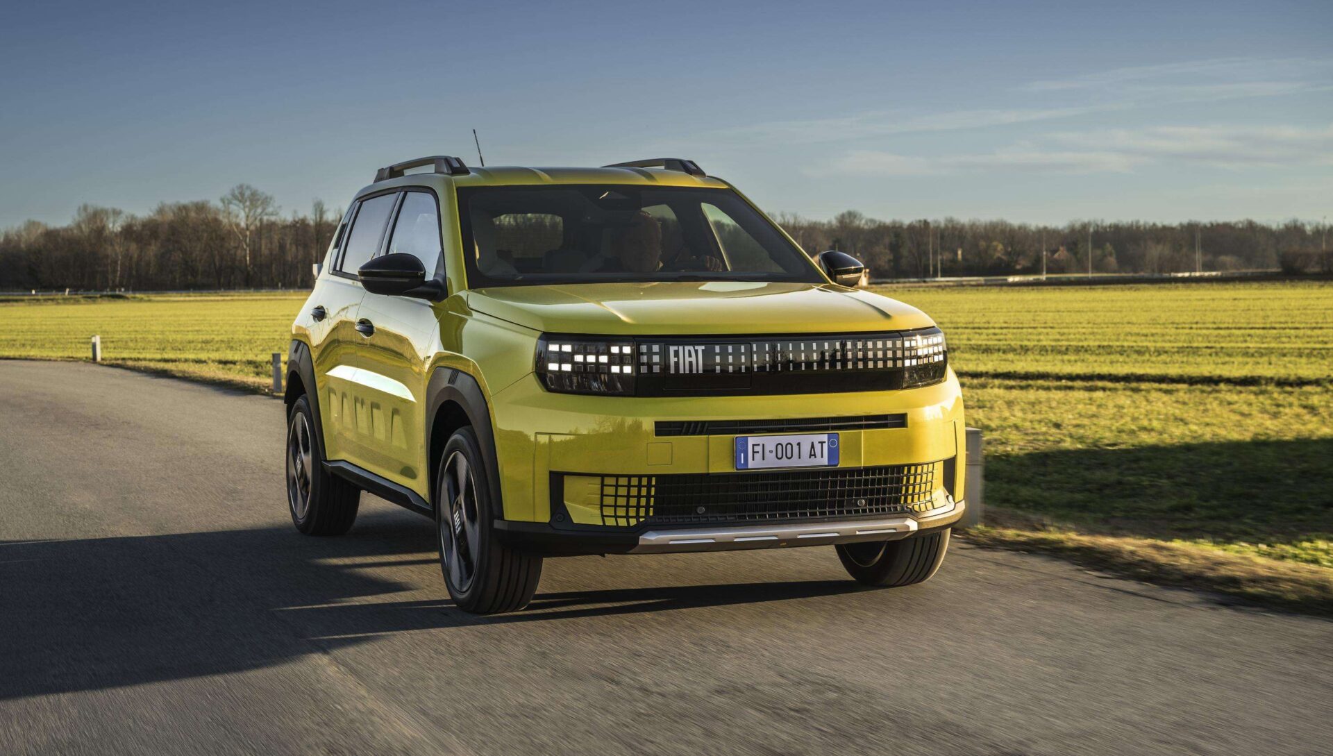 A bright yellow Fiat SUV driving on a paved road with a grassy field in the background.