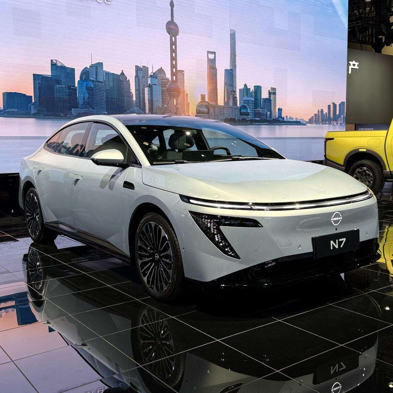 A sleek white electric car displayed at an auto show, featuring a modern design with distinctive headlights and stylish rims, set against a backdrop of the Shanghai skyline during sunset.