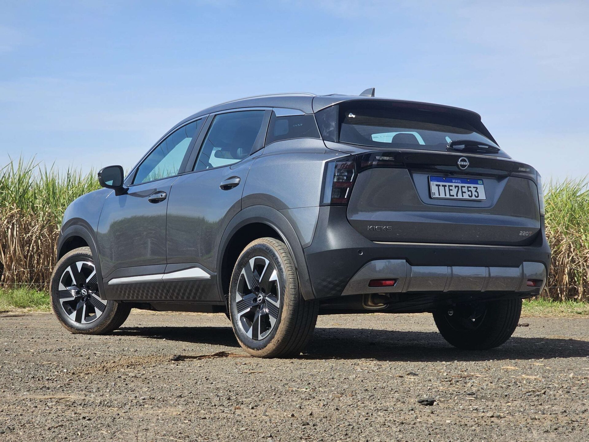 Vista lateral e traseira de um carro SUV Nissan Kicks, estacionado em uma estrada de cascalho com canaviais ao fundo sob um céu claro.