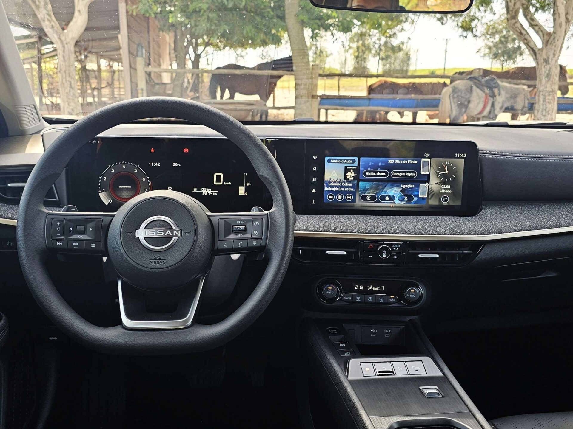 Painel interno de um carro Nissan, com volante, tela digital mostrando informações e sistema de entretenimento no console central.