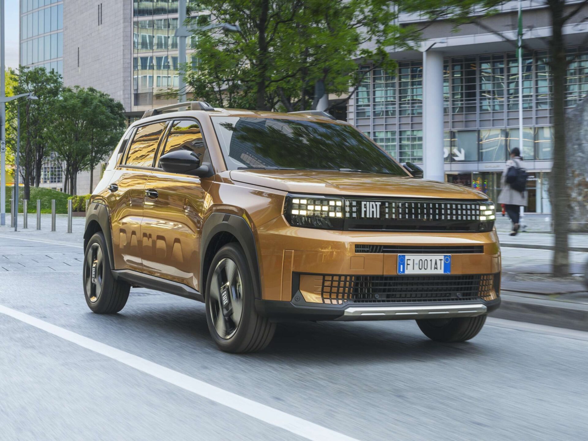 A bronze Fiat Panda driving on a city street with modern buildings and green trees in the background.