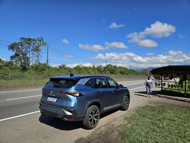 A blue SUV parked on the side of a road with a person standing nearby, under a shaded area, surrounded by greenery and a clear blue sky.