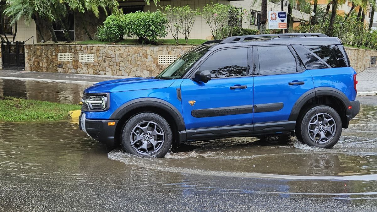 A blue SUV driving through a flooded street, partially submerged in water with reflections visible on the surface.