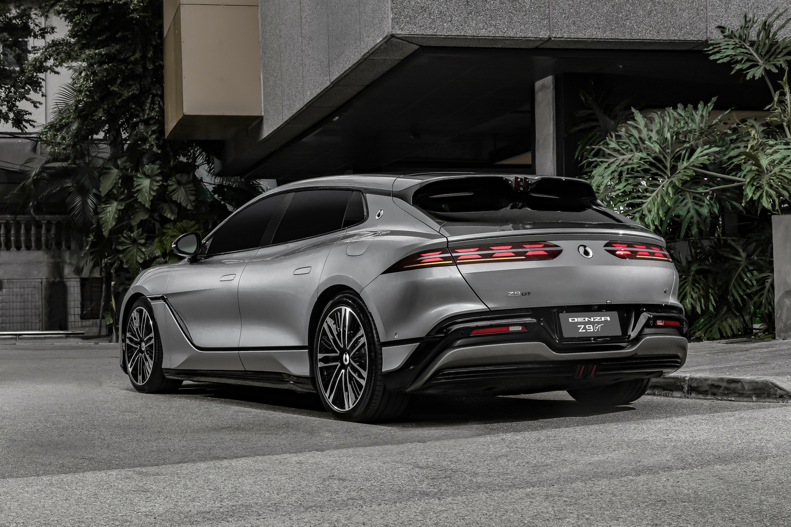 A sleek silver Denza Z9 GT parked on a street, showcasing its modern design and distinctive taillights, surrounded by greenery and urban architecture.