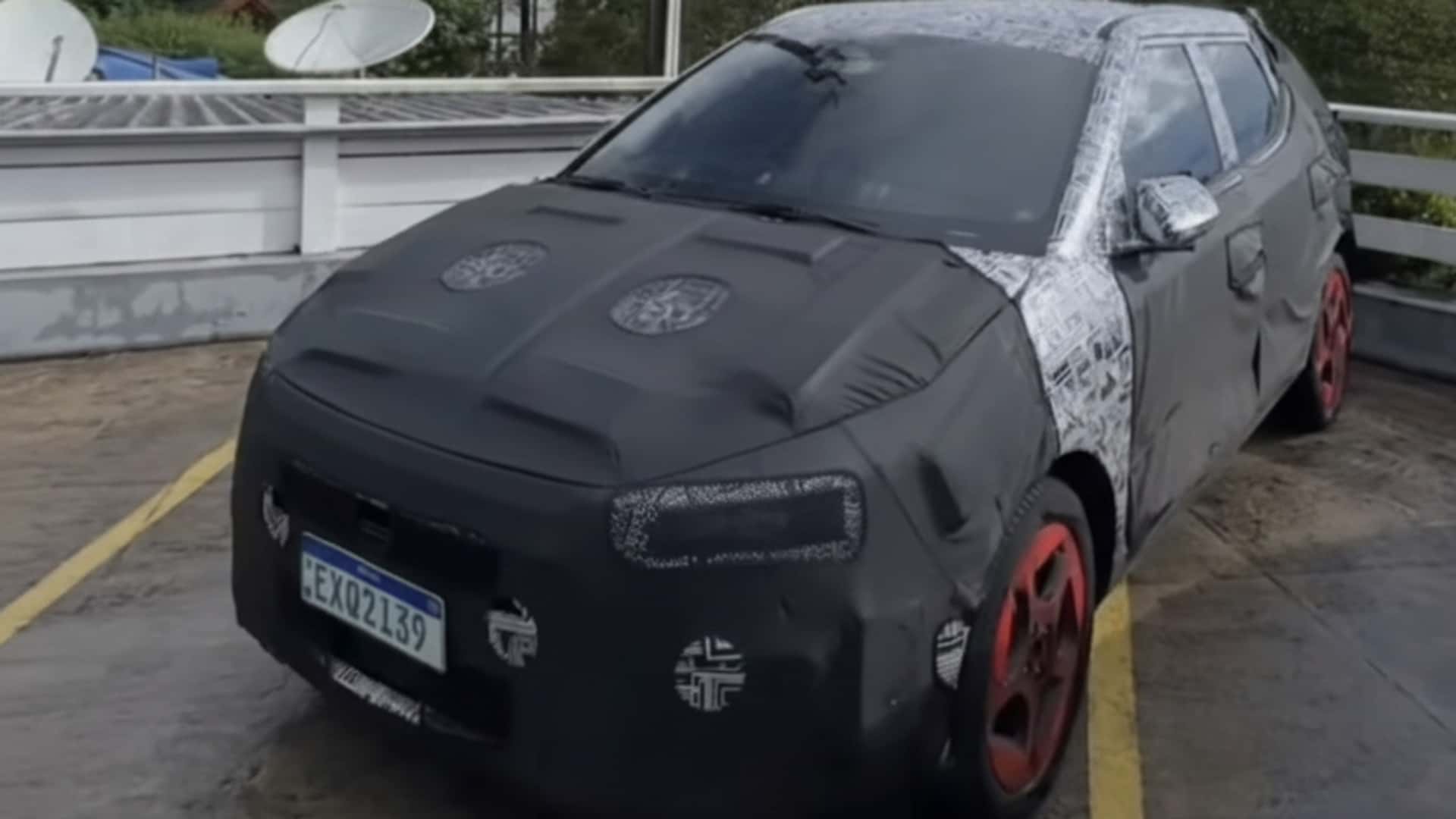 A camouflaged car covered in black and white patterns, parked on a concrete surface, with red wheels and satellite dishes visible in the background.