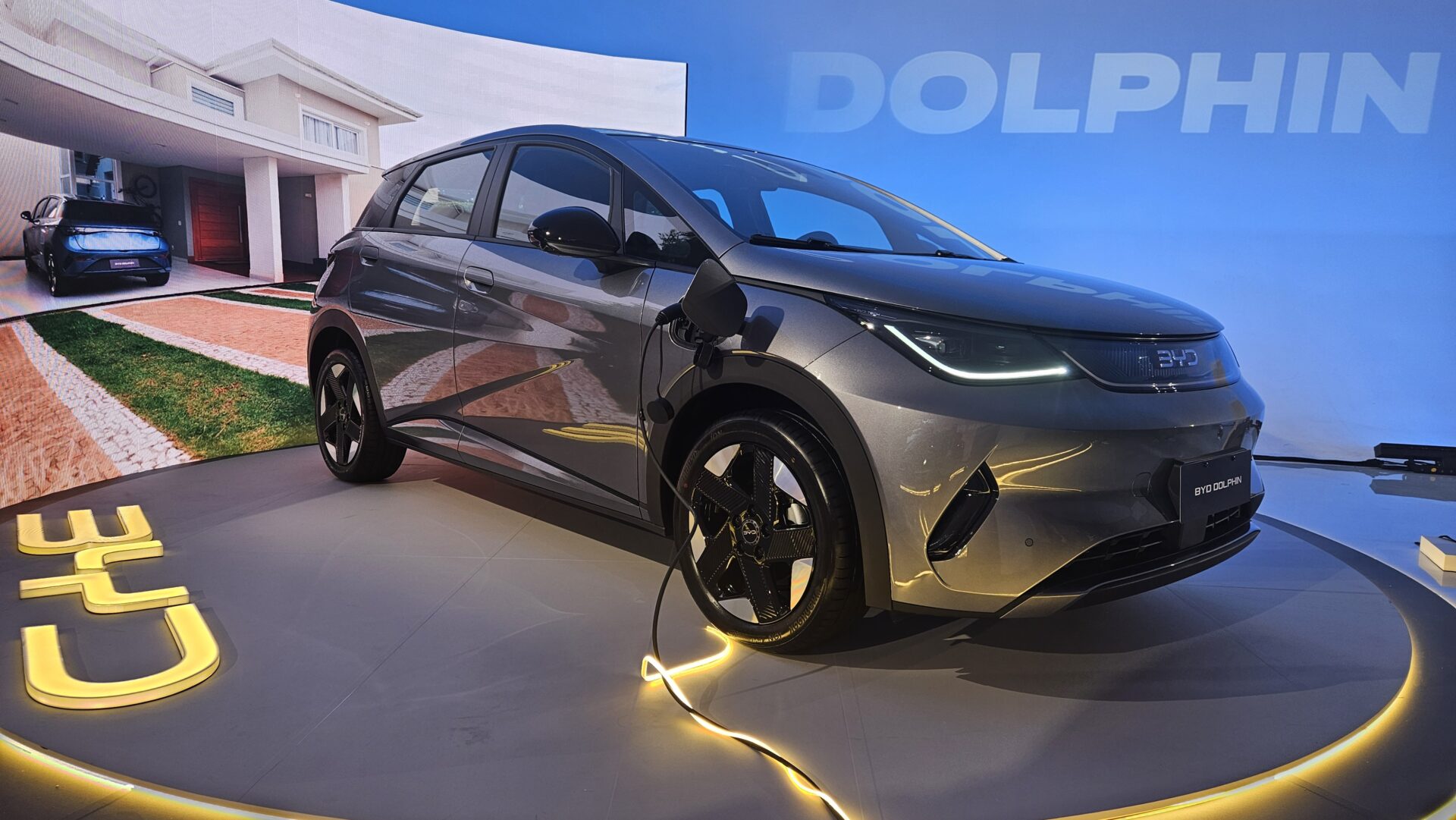 Carro elétrico BYD Dolphin em exibição, com um design moderno e pintura metálica cinza. A imagem de fundo exibe uma casa com um carro estacionado. O carro está conectado a um carregador elétrico.