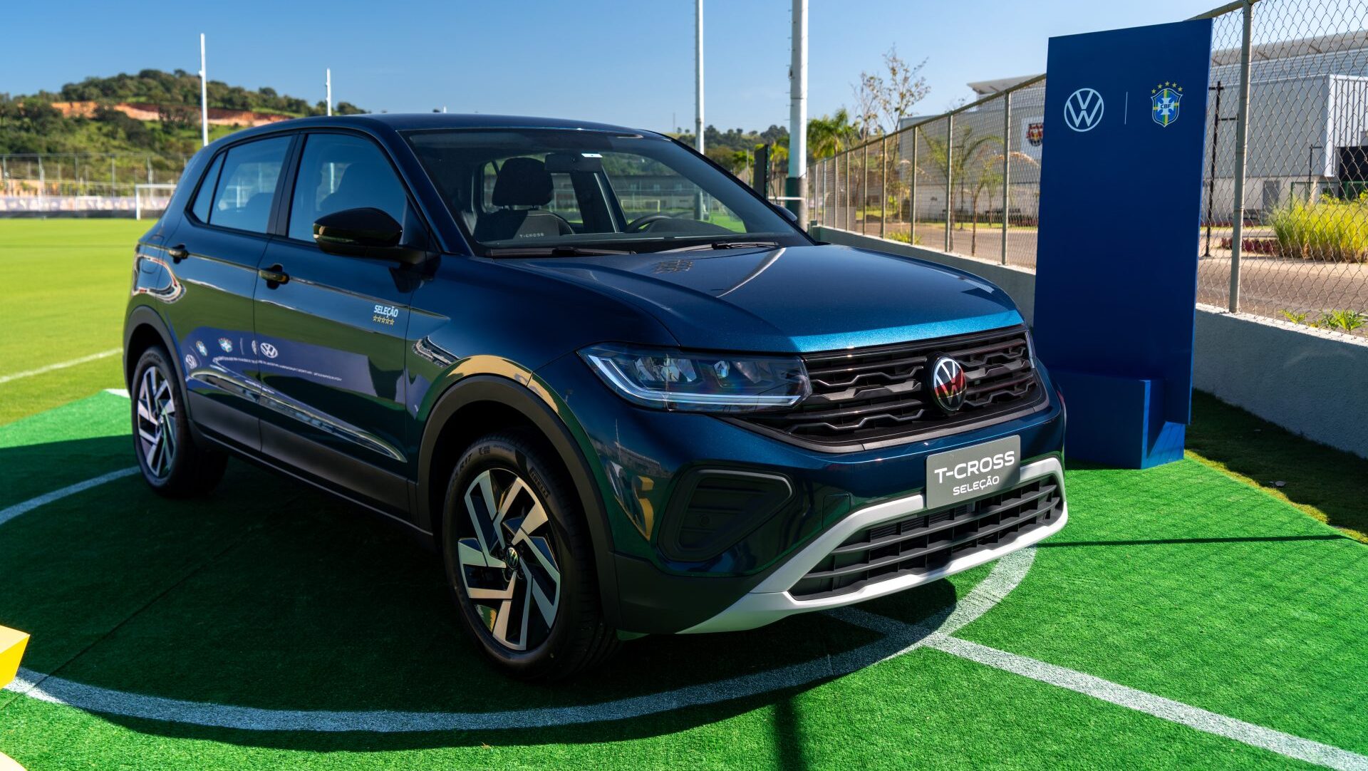 Carro Volkswagen T-Cross em um campo de futebol, com detalhes em azul e branco, e placa 'Seleção' ao lado.