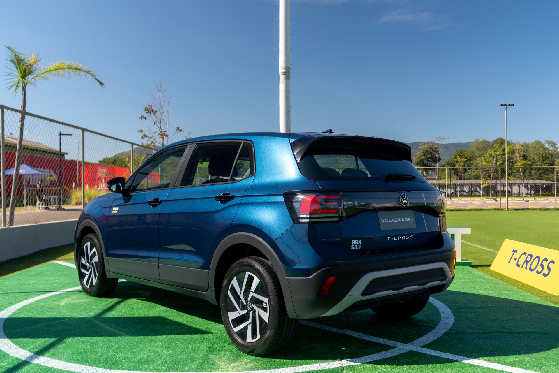 Carro Volkswagen T-Cross azul estacionado em um campo de grama sintética, com cercas ao fundo e céu claro.