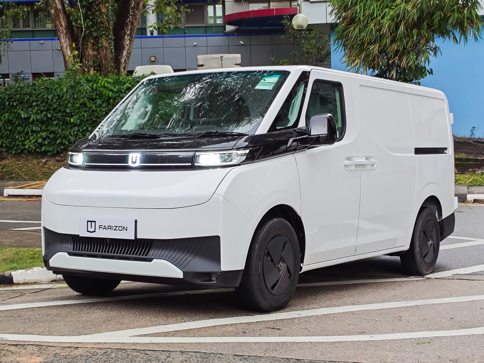 A modern white delivery van with a black front hood parked on a street, surrounded by greenery and a building in the background.