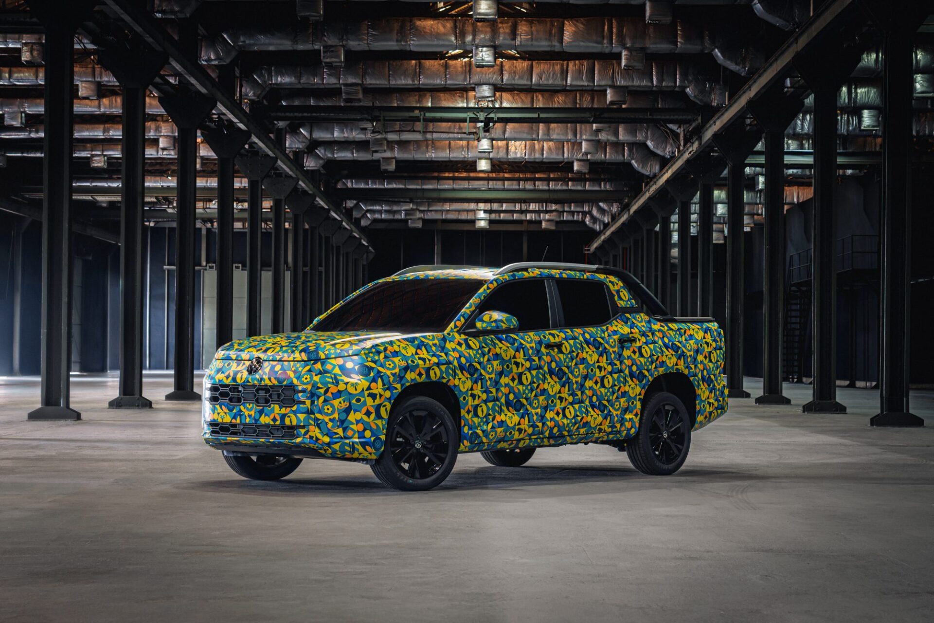 Um carro coberto com uma embalagem colorida de flores em um grande espaço industrial com colunas pretas.