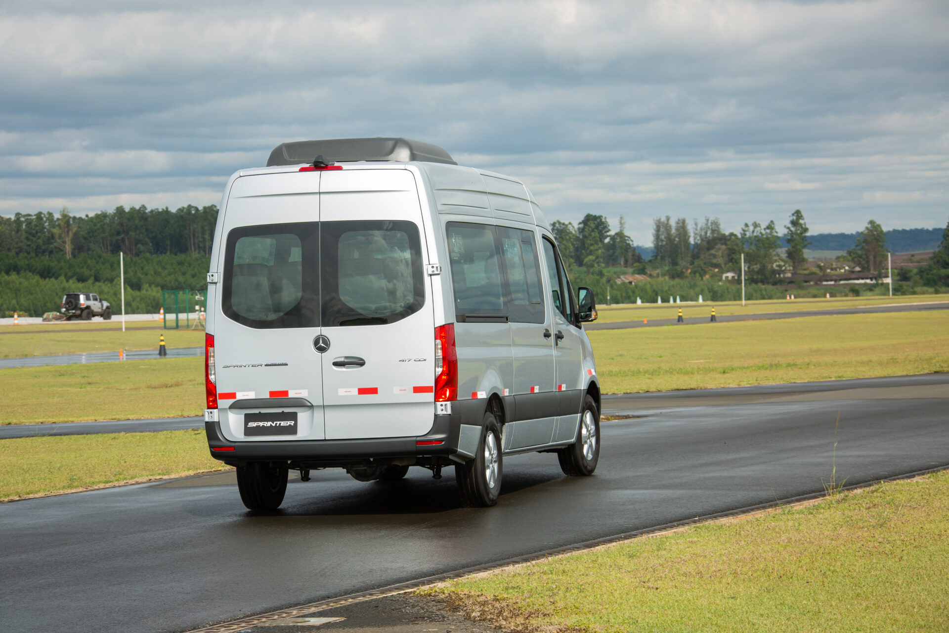 Van Mercedes-Benz Sprinter em uma pista, vista traseira, com céu nublado ao fundo e área verde ao redor.