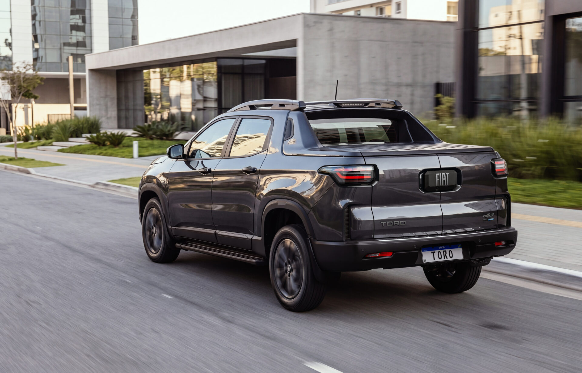 Um carro utilitário esportivo Fiat Toro cinza se deslocando na rua, com edifícios modernos ao fundo.
