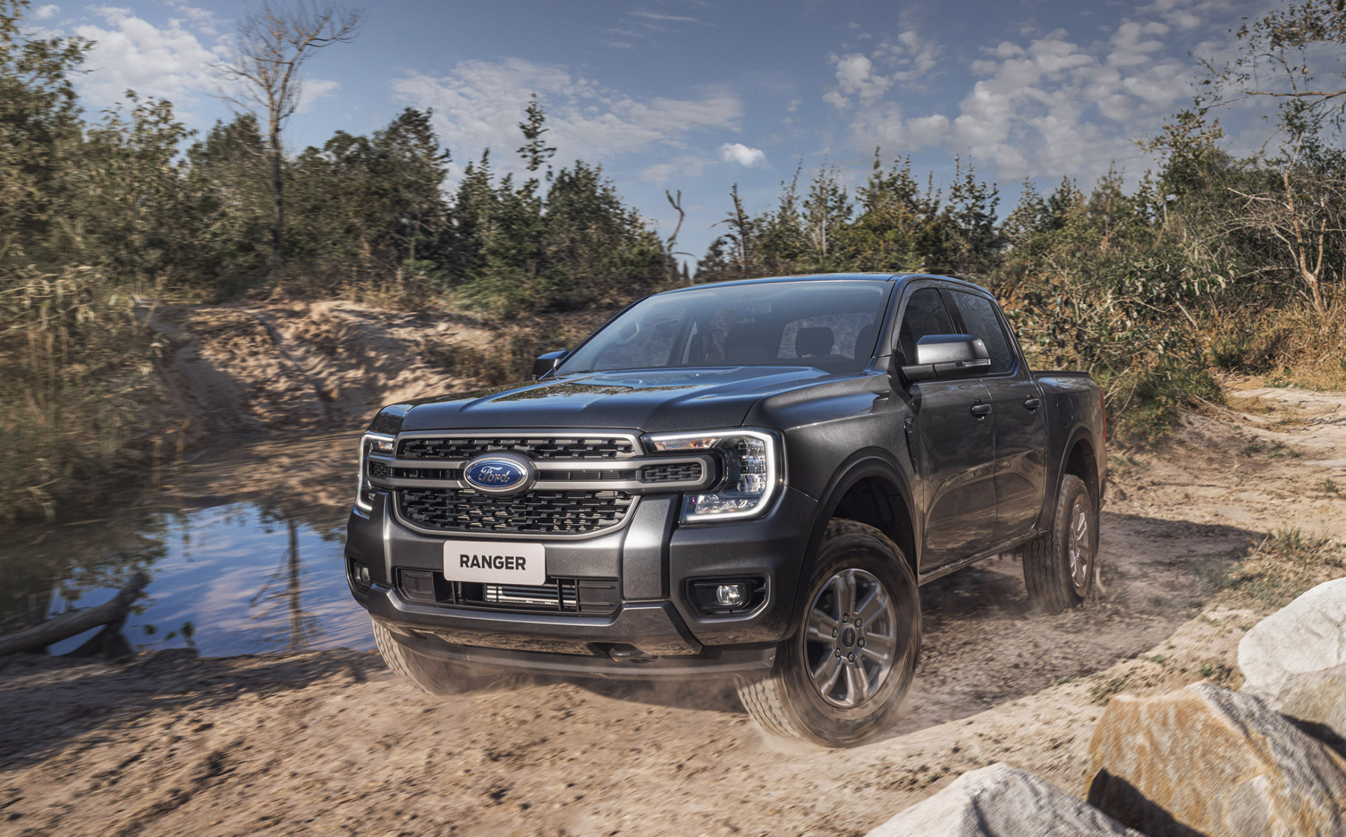 Uma caminhonete Ford Ranger em um terreno off-road, com vegetação ao fundo e céu azul.