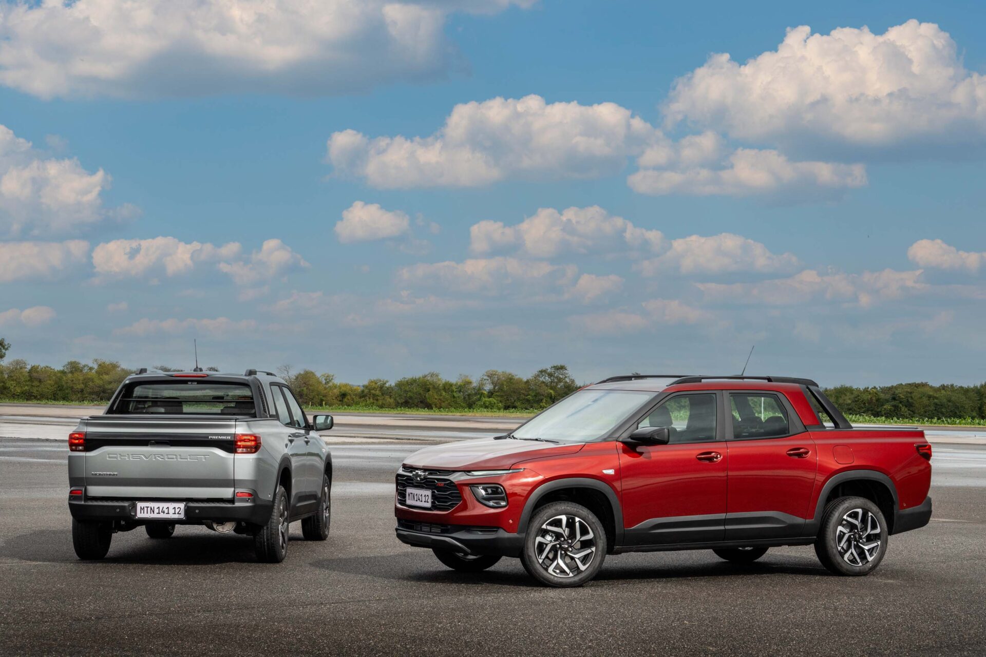 Two Chevrolet vehicles parked on a paved surface under a blue sky with clouds. The left vehicle is a grey model, viewed from the rear, and the right vehicle is a red model, viewed from the front.