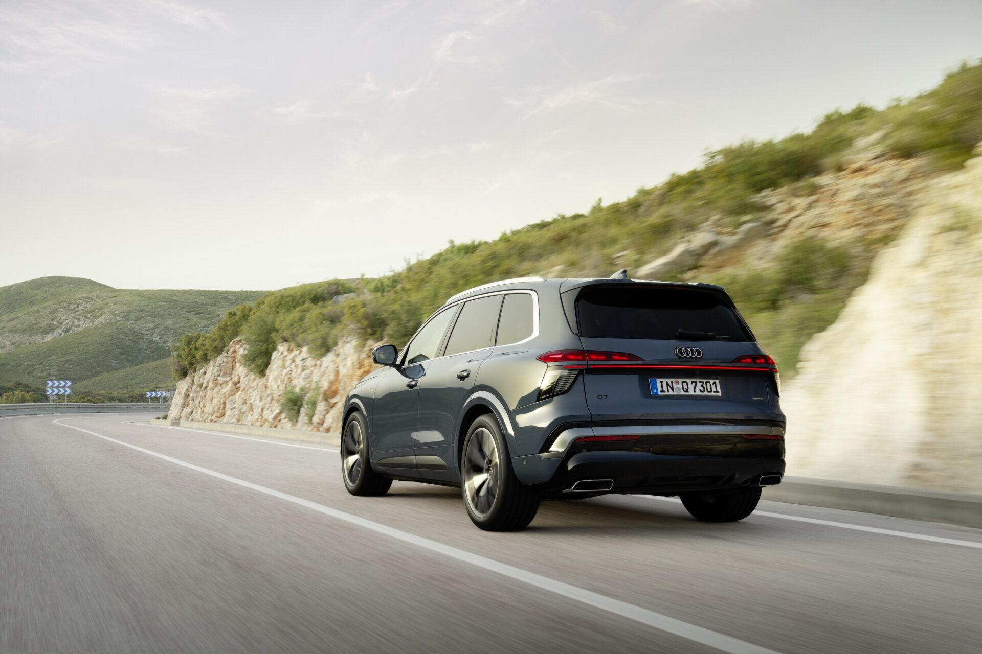 A blue Audi Q7 SUV driving along a winding road surrounded by lush green hills and rocky cliffs.