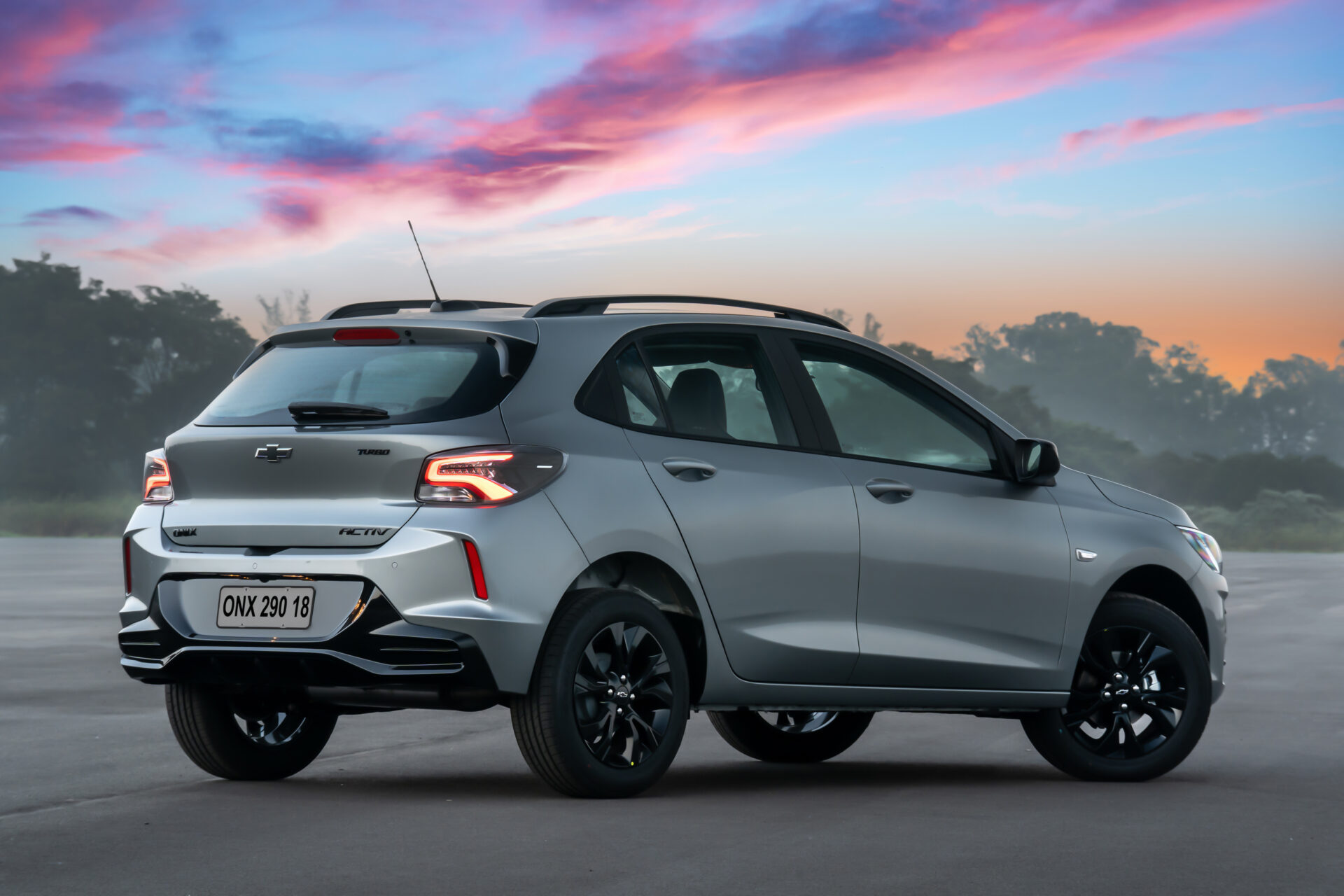 A silver Chevrolet car, model Onix Activ, parked on pavement with a scenic sunset background featuring pink and orange hues.