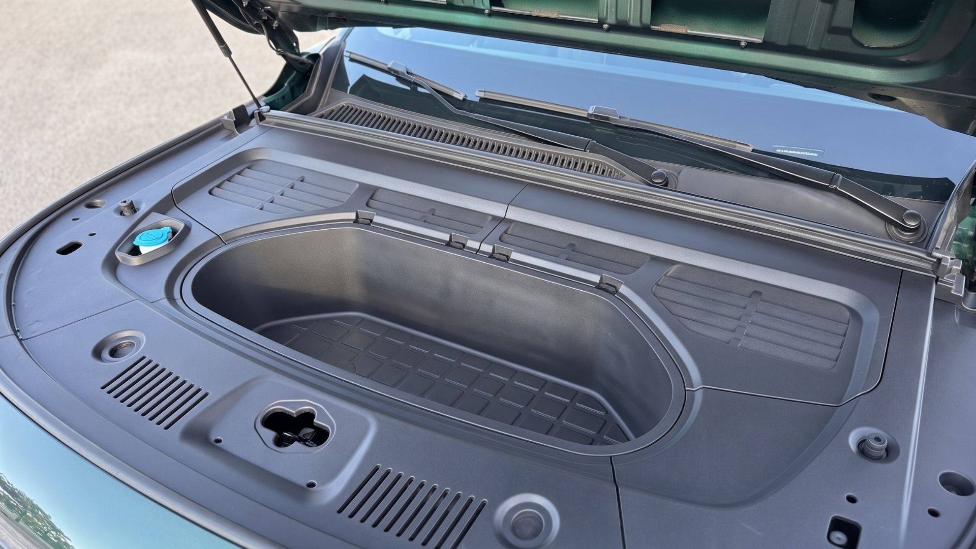 An overhead view of the inside of a car hood, showing a large, open compartment with a textured bottom. Various attachments and fittings are visible, including a blue reservoir cap and ventilation slots.