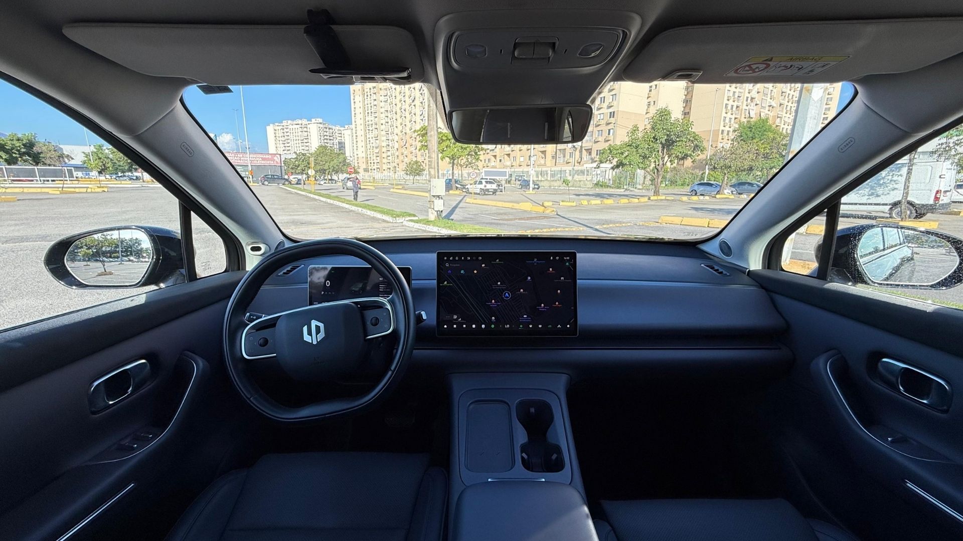 Interior view of a modern car showcasing a steering wheel, a large digital display panel, and a sleek dashboard design, with a view of a parking area and buildings outside the windows.
