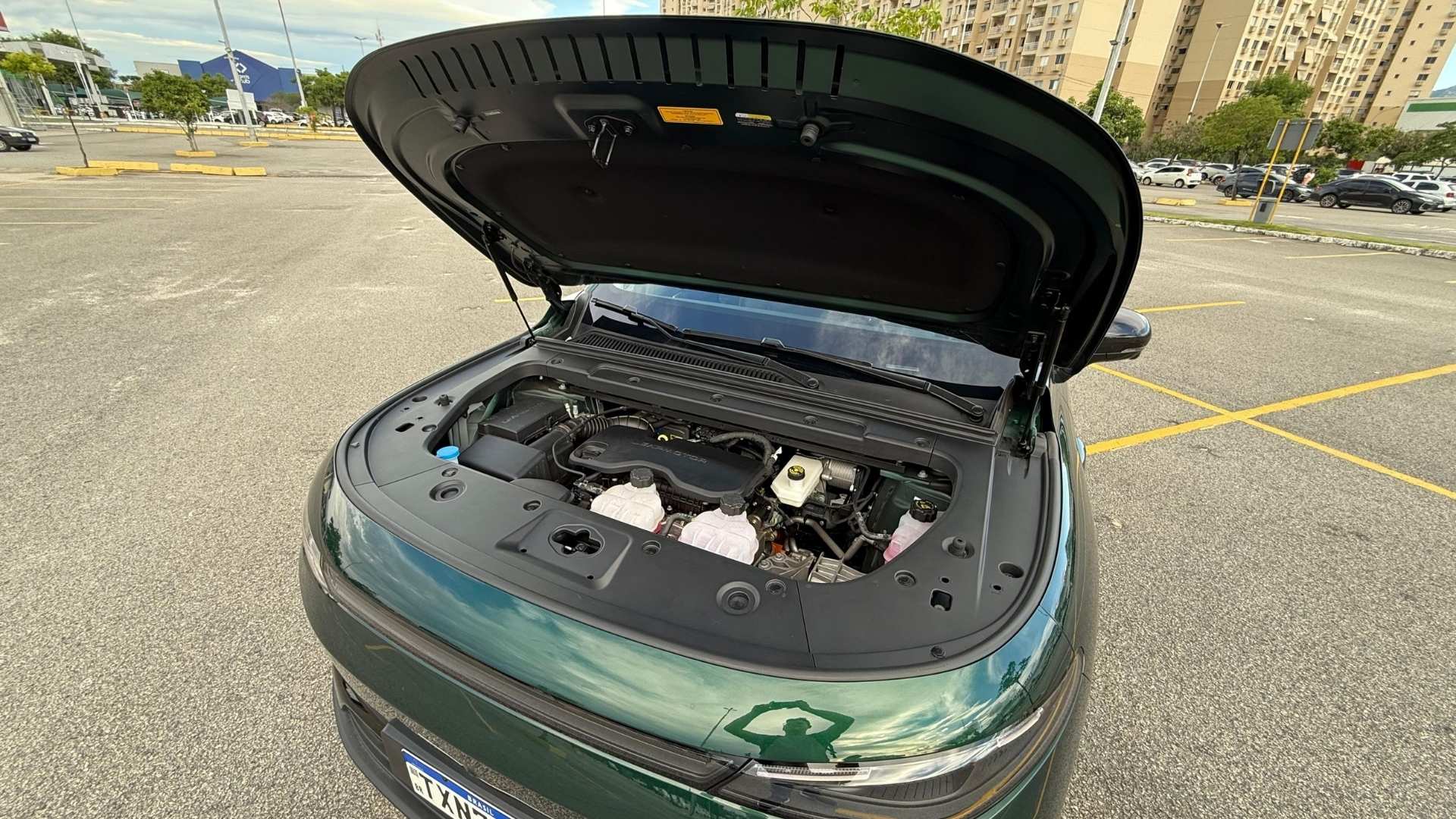 Image of an open car hood revealing the engine compartment of a green vehicle, parked in an empty lot with buildings in the background.