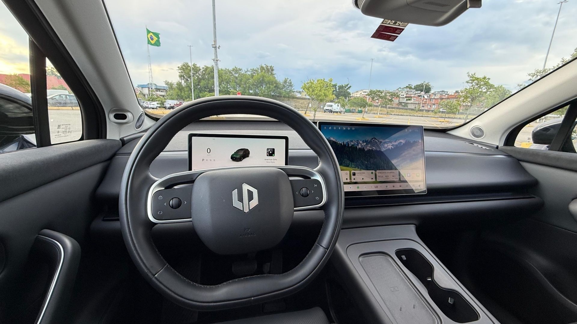 Interior view of a modern car featuring a steering wheel with a logo, a digital display panel showing the vehicle's status, and a secondary screen displaying a landscape. A Brazilian flag is visible in the background.