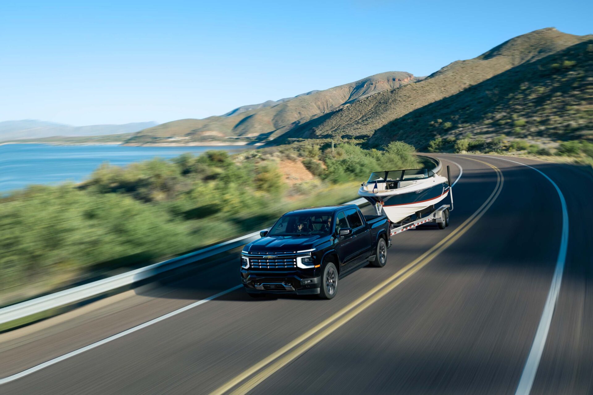 A black pickup truck towing a boat on a winding road with a scenic view of a lake and mountains in the background.