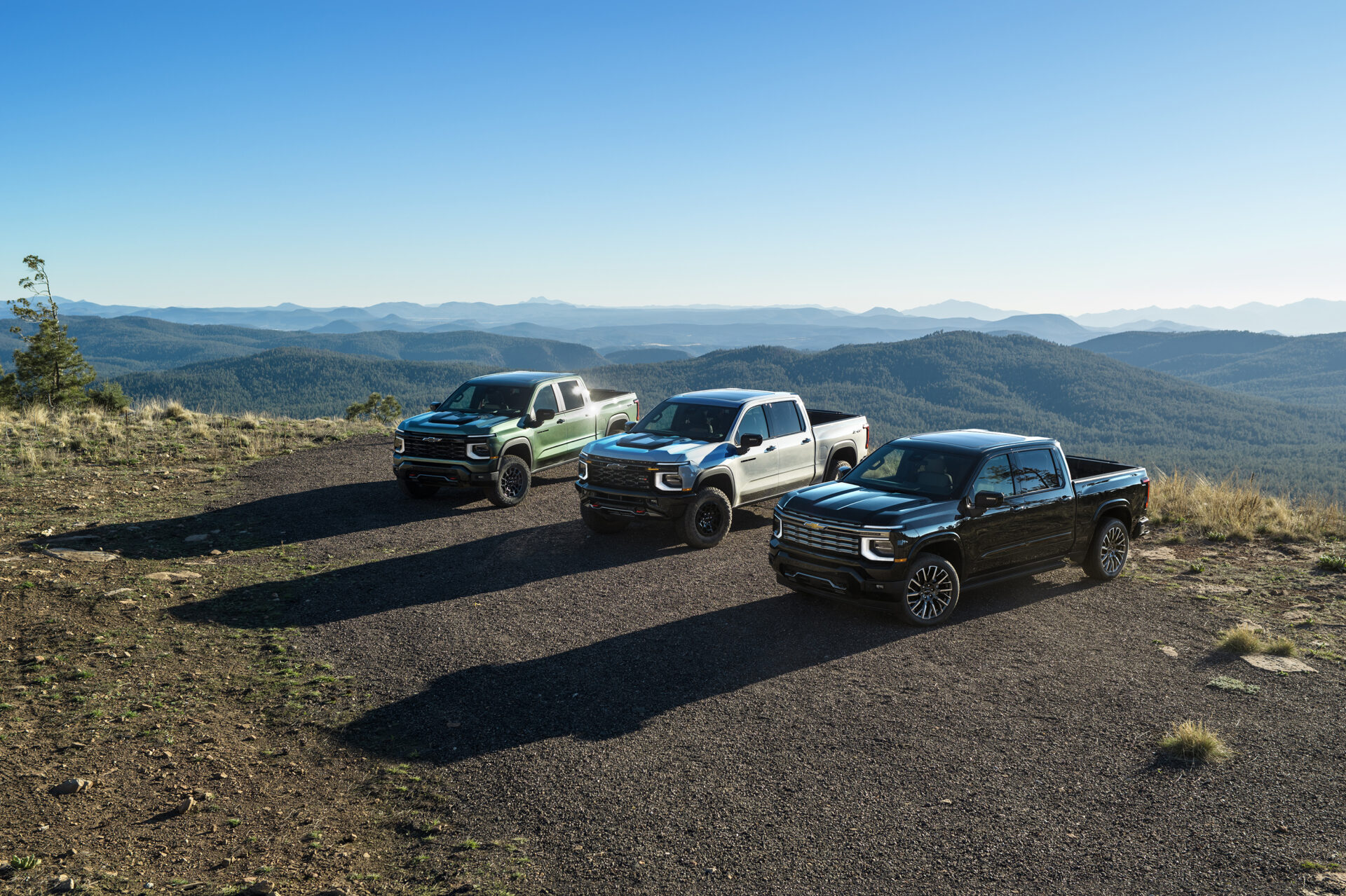 Three pickup trucks parked on a gravel area with a mountainous landscape in the background, under a clear blue sky.