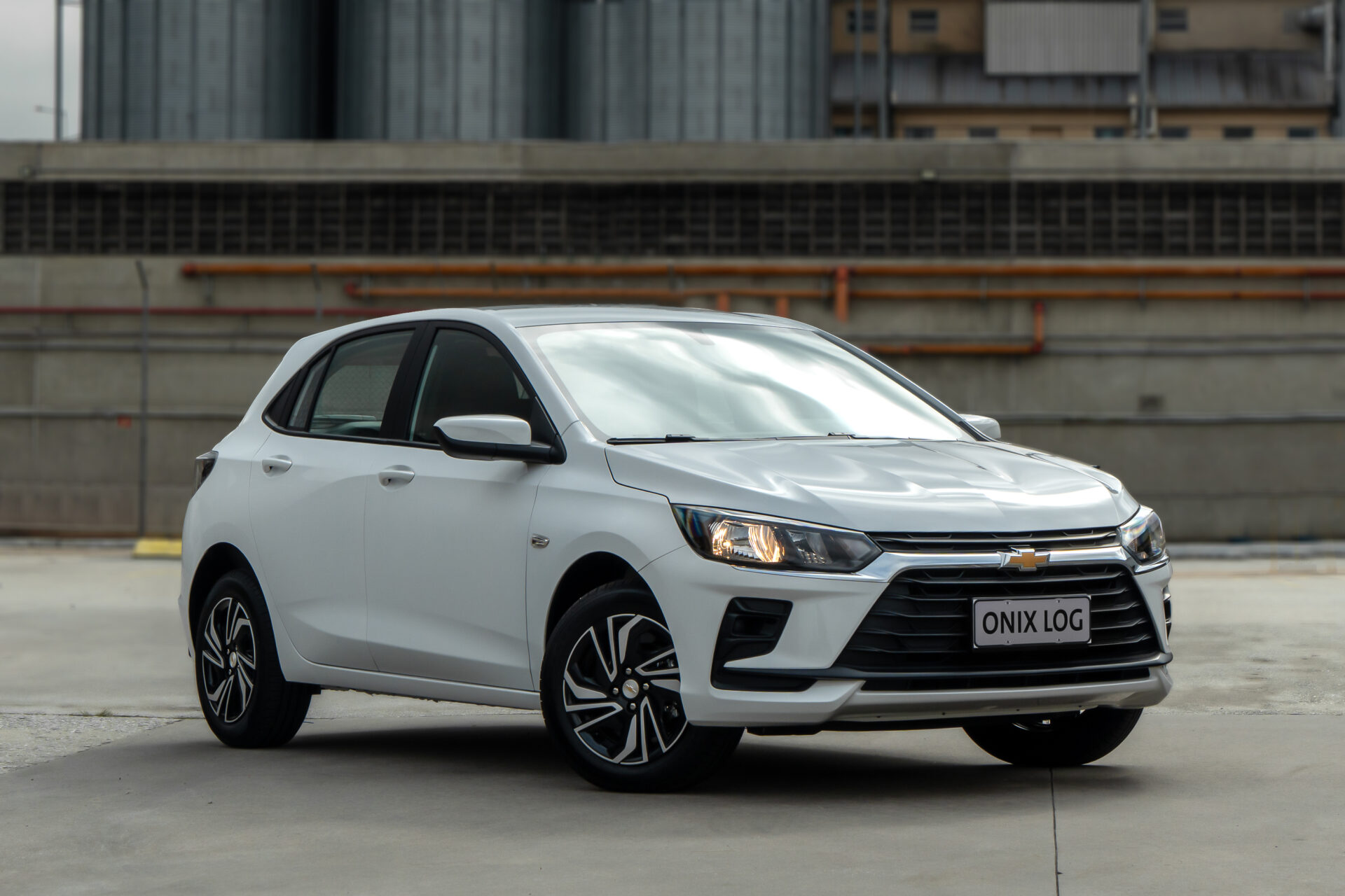 A silver Chevrolet Onix hatchback parked in an industrial area with a modern building in the background.