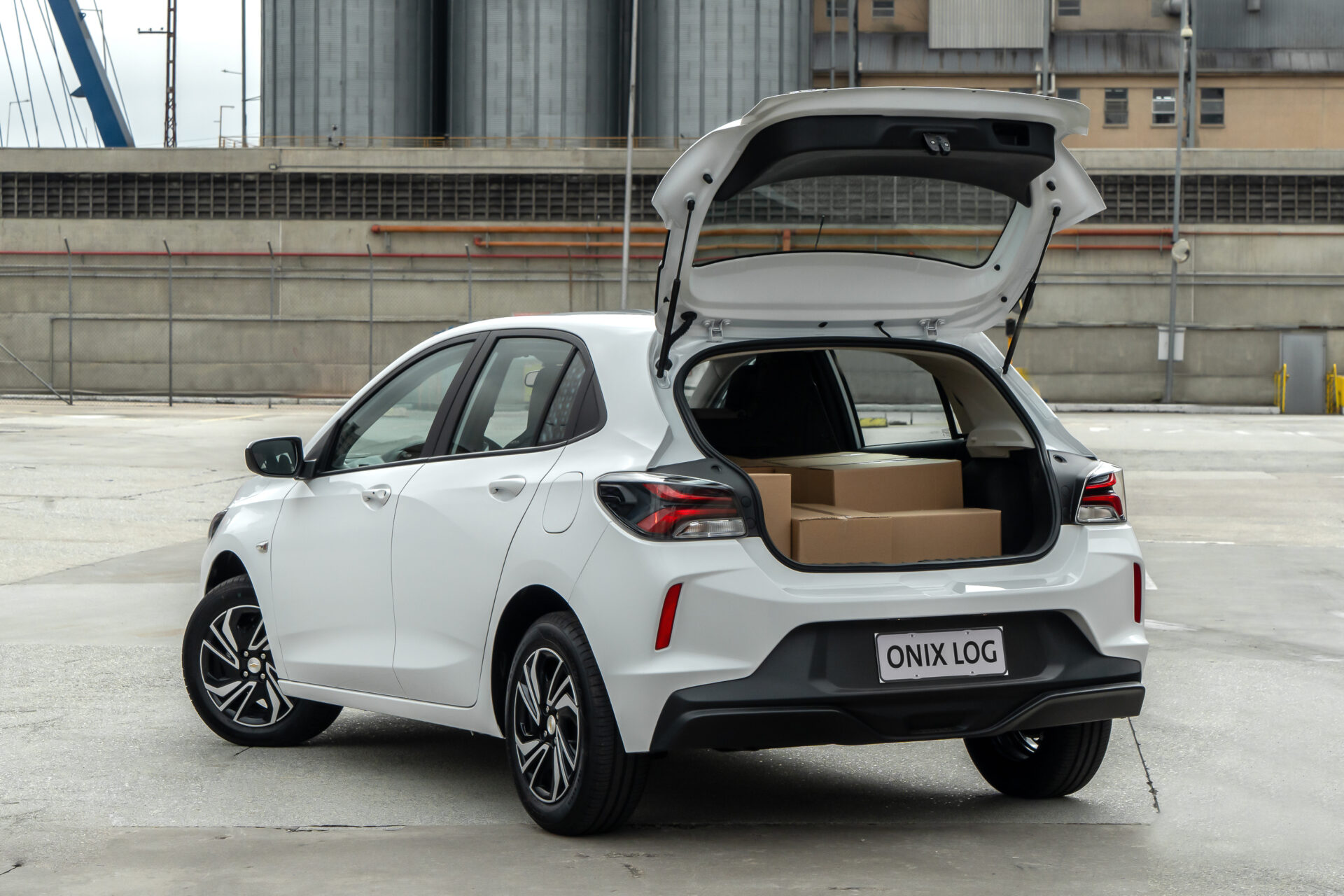 A white hatchback car parked with its rear hatch open, revealing cardboard boxes inside the trunk in an industrial setting.