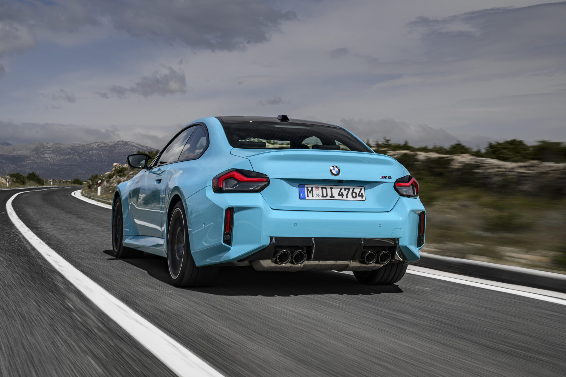 A vibrant blue BMW M2 driving on a winding road with mountains in the background.