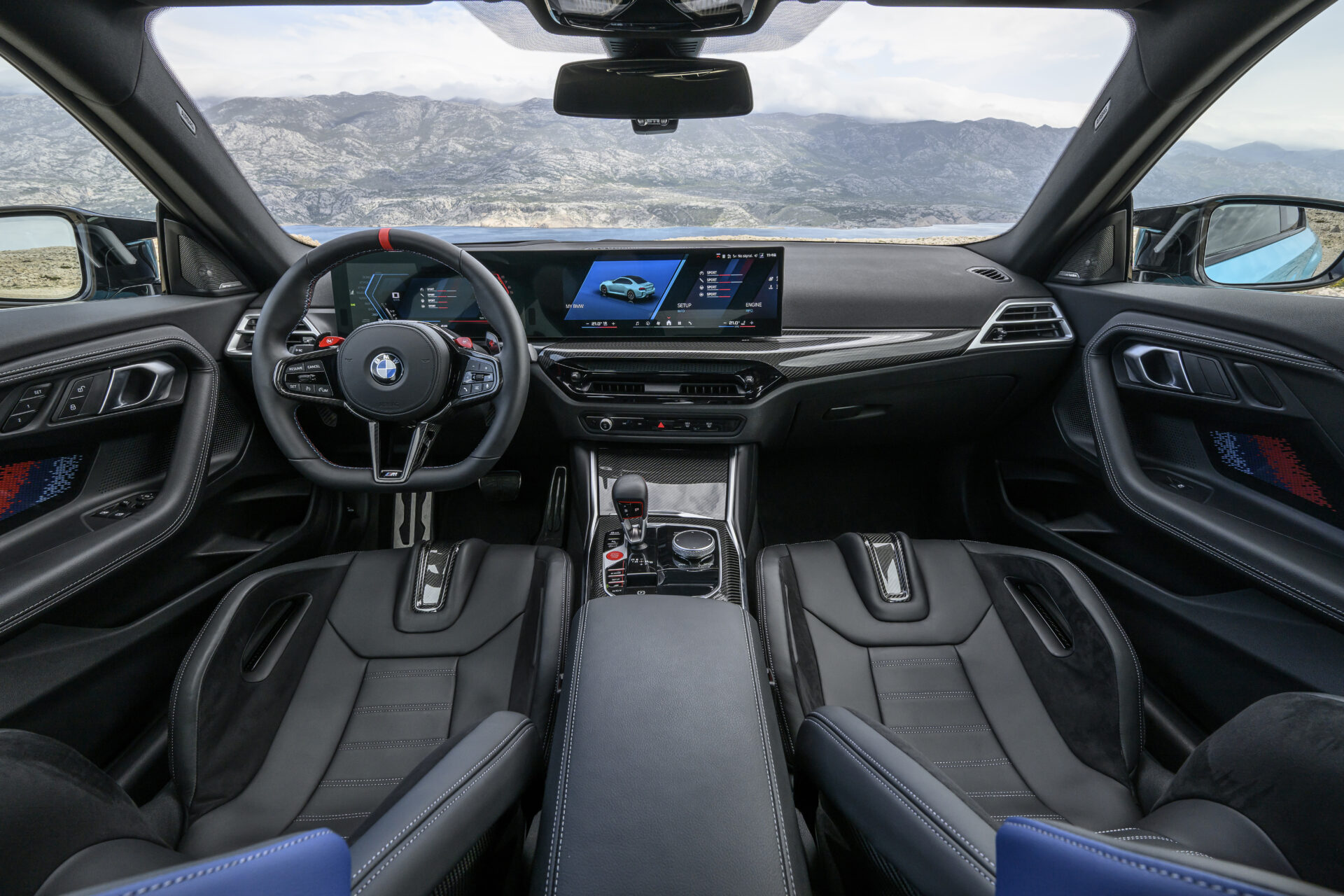 Interior view of a modern BMW car showcasing a sleek dashboard, sporty steering wheel, advanced digital display, and leather seats with a panoramic view of mountains in the background.