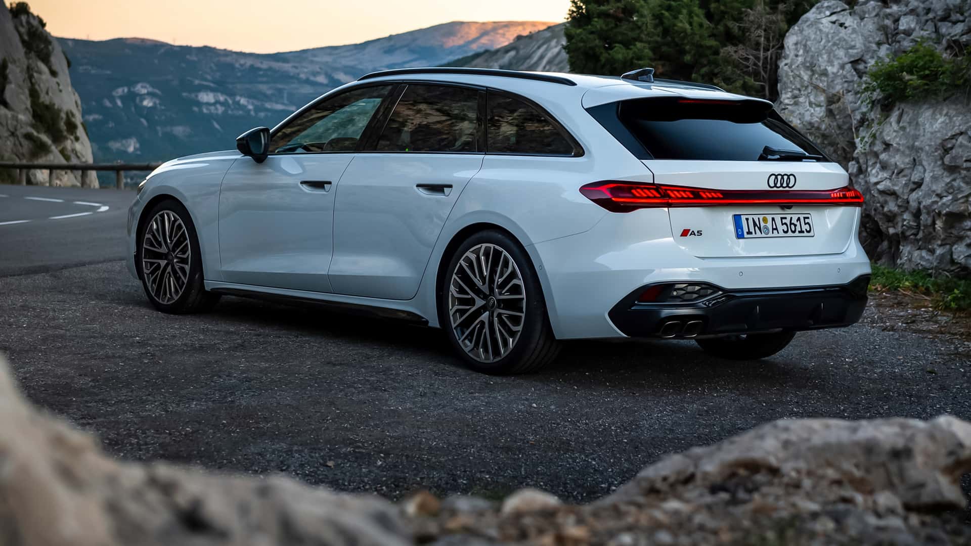 A white Audi A5 Sportback parked on a scenic mountain road with rocky terrain and a panoramic view in the background.