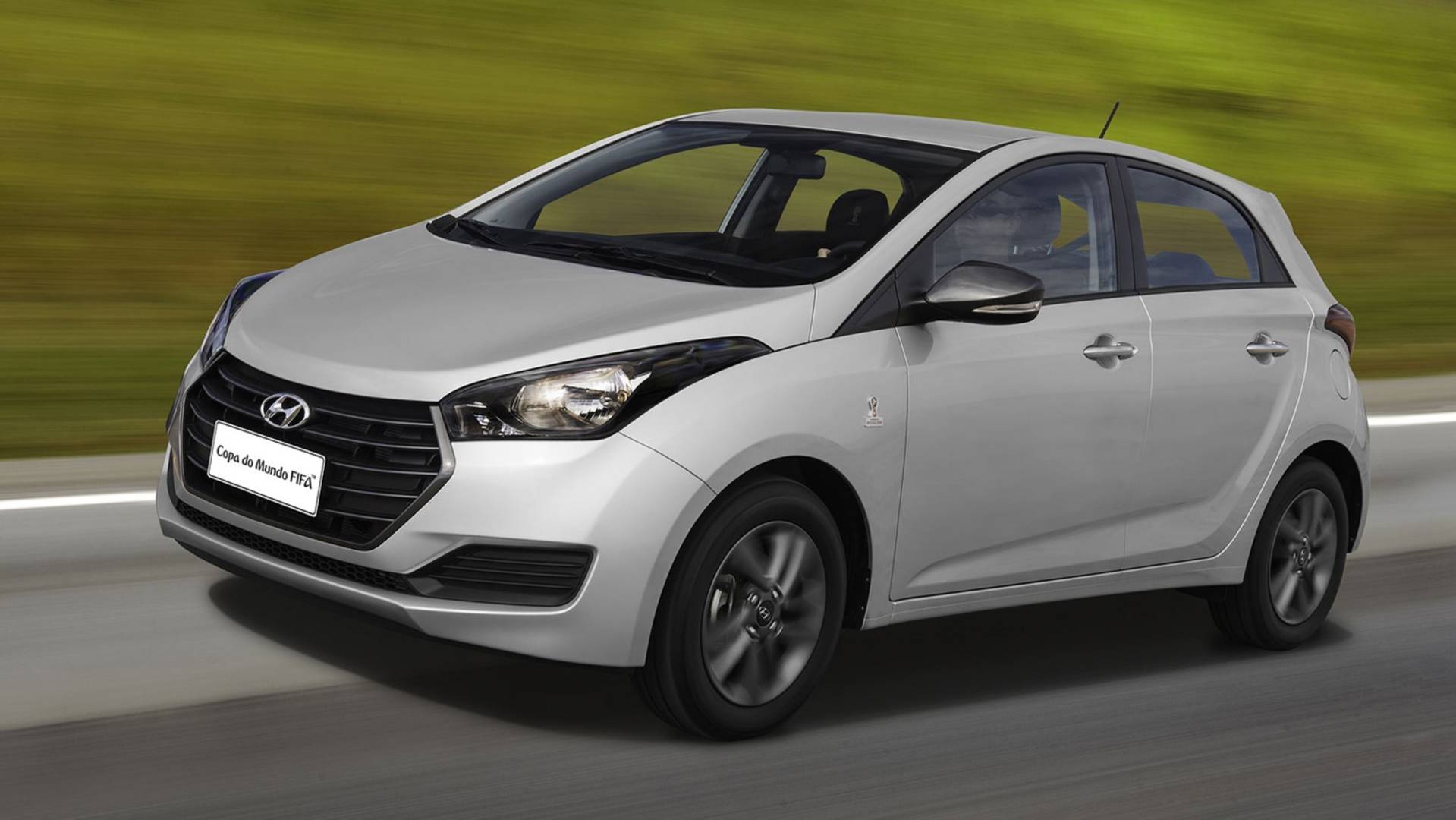 A silver Hyundai hatchback car driving on a road with a blurred green background, showcasing its sleek design and modern features.