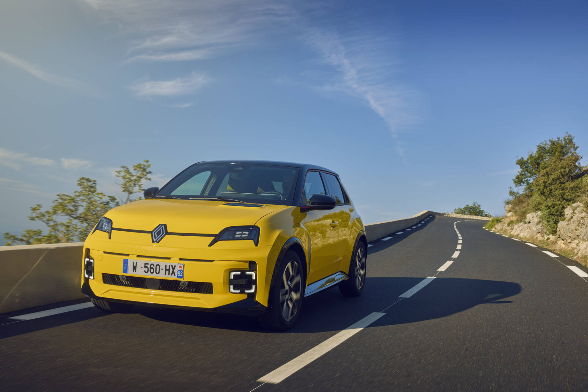A bright yellow Renault car driving along a winding mountain road under a clear blue sky.