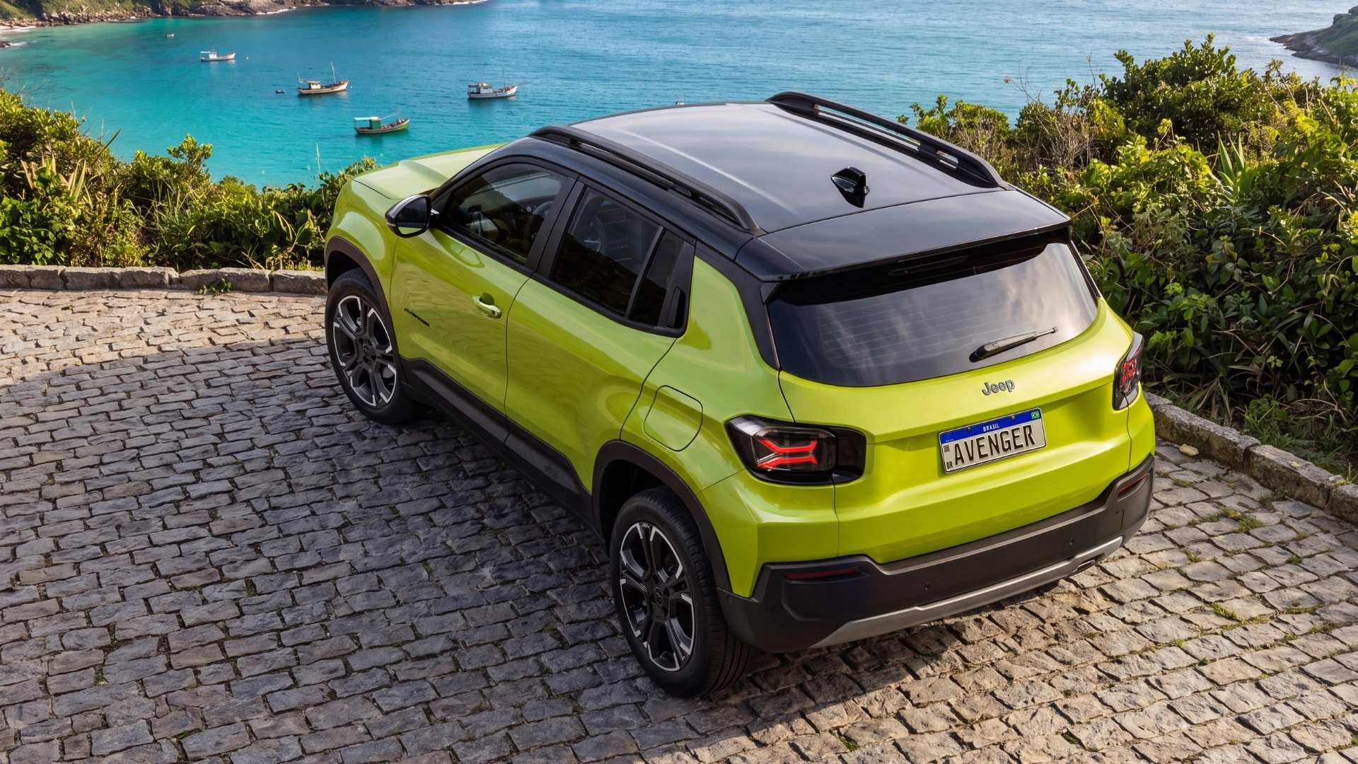 A vibrant green Jeep Avenger parked on a cobblestone path overlooking a blue ocean with boats in the distance.
