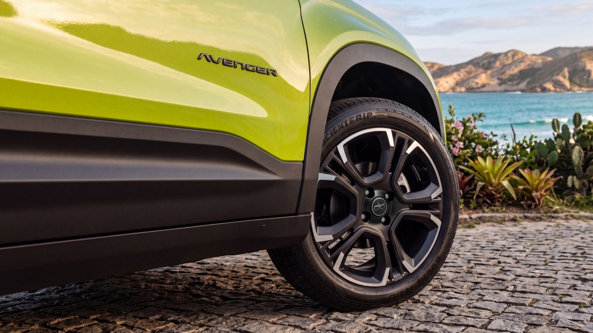 Close-up of a green car with the word 'Avenger' visible, showcasing its front wheel and alloy rim against a scenic beach background.