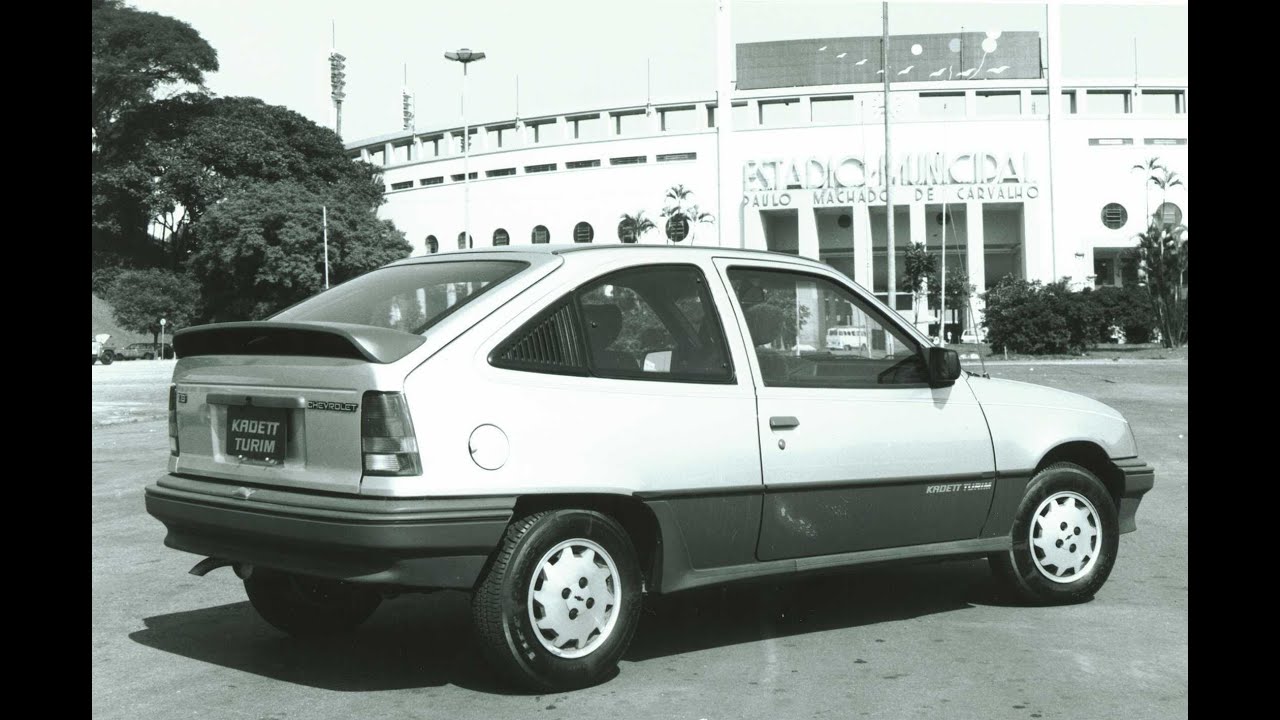 A black and white photograph of a Chevrolet Kadett Turim parked in front of a large stadium, identified as Estádio Municipal Paulo Machado de Carvalho, with trees and poles visible in the background.