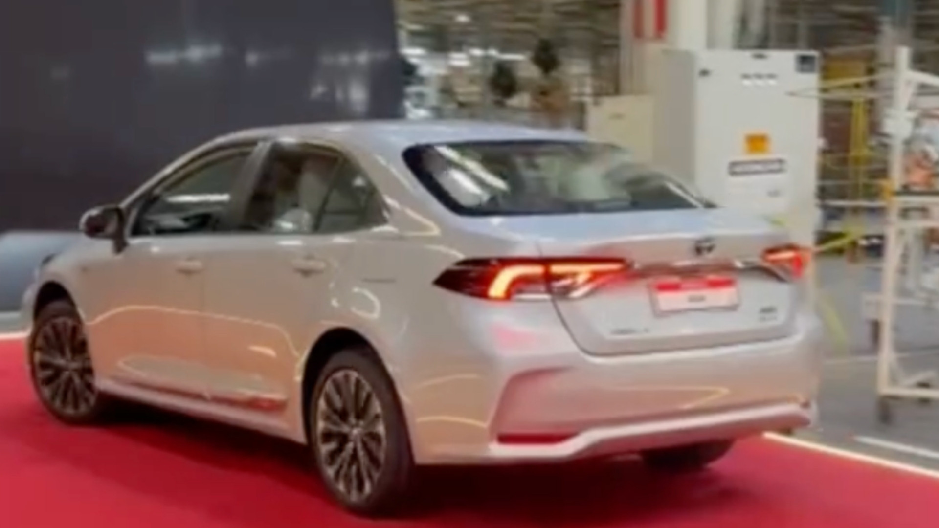 A rear view of a silver sedan parked on a red carpet inside a large industrial setting, showcasing its modern design and alloy wheels.