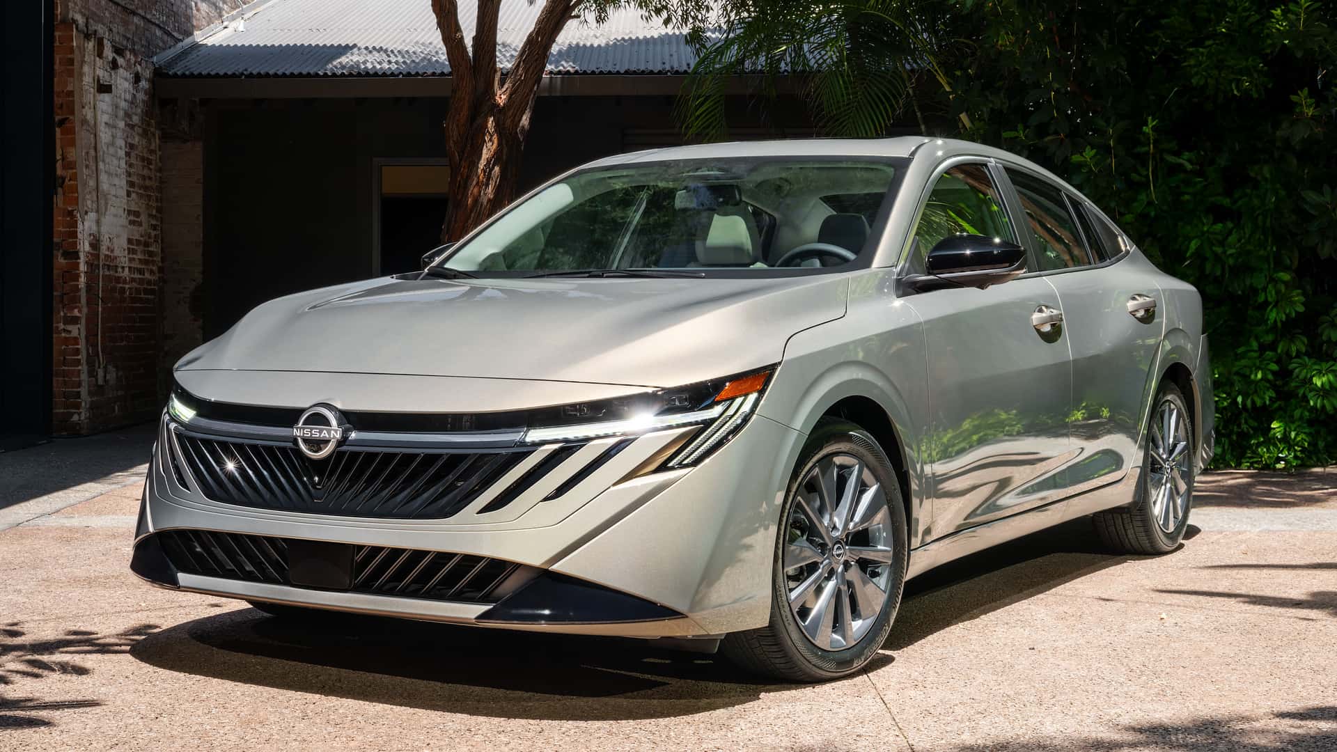 A silver Nissan sedan parked in a sunlit outdoor setting, showcasing its modern design and sleek lines.