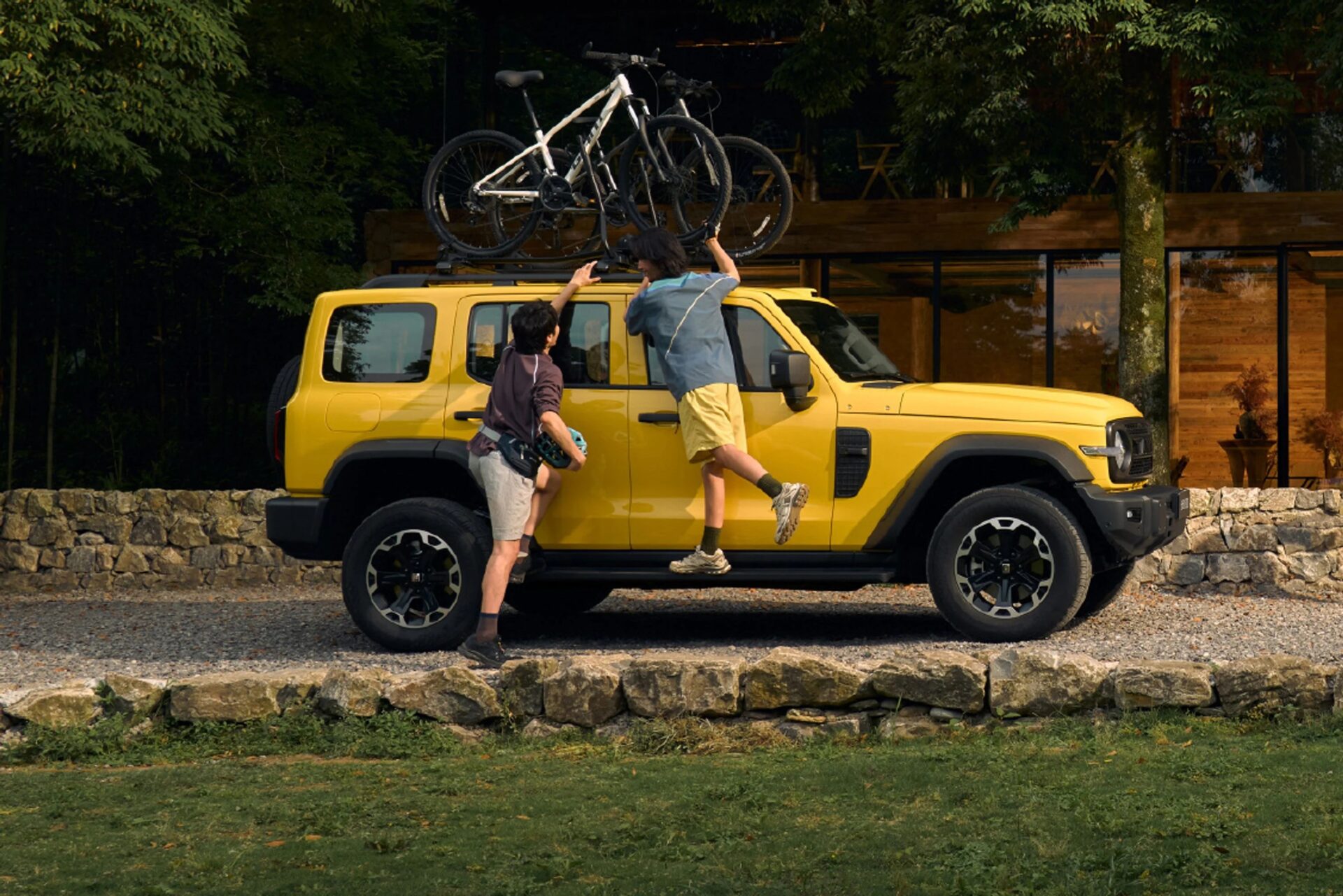 A yellow SUV parked in a scenic area with two people loading bicycles onto its roof rack, one person climbing and the other assisting.