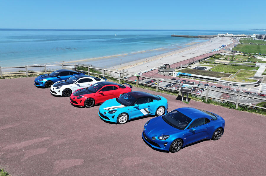 A lineup of five colorful sports cars parked on a scenic overlook, with a beach and coastline in the background on a clear, sunny day.