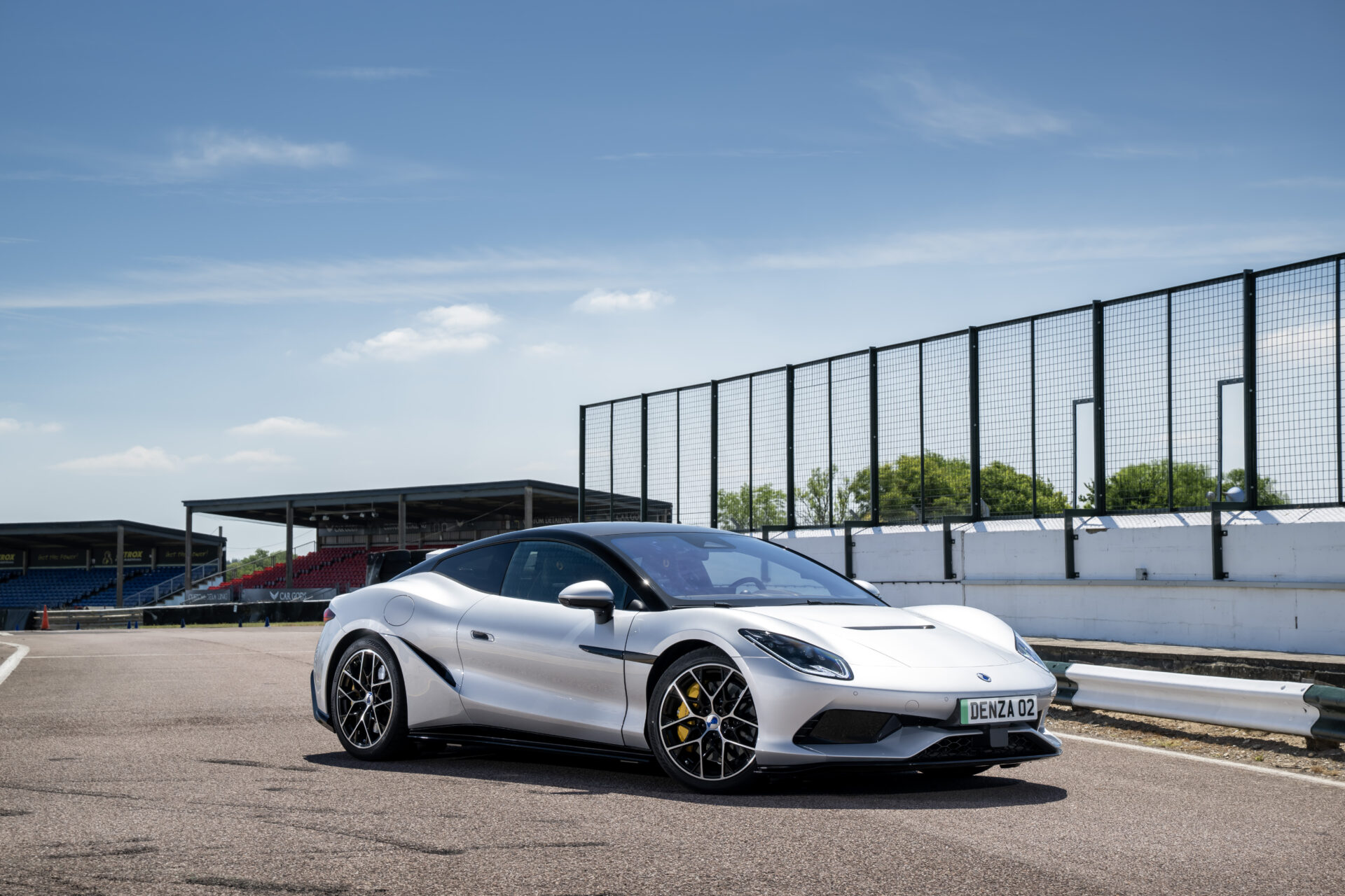 A sleek silver sports car parked on a racetrack, with bright blue skies and green trees in the background.